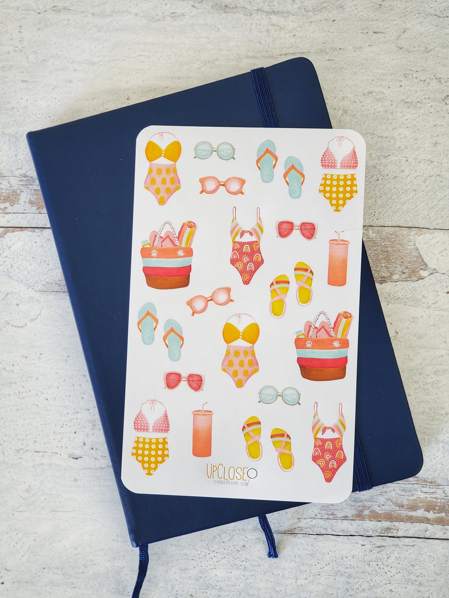 This sticker sheet features colorful illustrations of beach day essentials, including bathing suits, flip flops, sunglasses, a beach bag and a tumbler. 