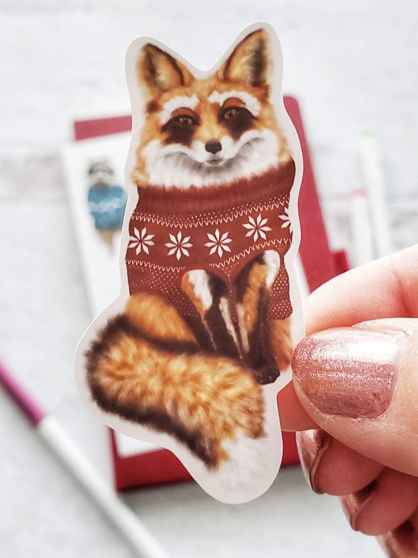 Close up of an illustrated fox sticker. The fox is wearing a red sweater with white snowflakes and polka dots.