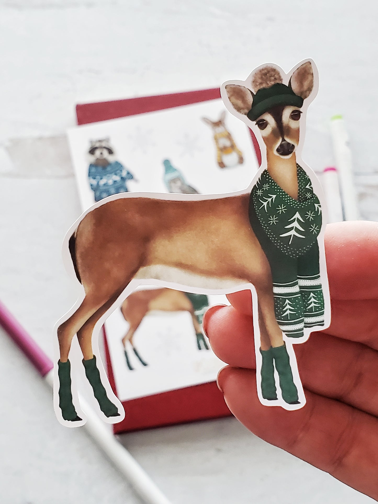 Close up of a sticker of an illustrated deer wearing a green stocking hat and matching scarf and leg warmers.  