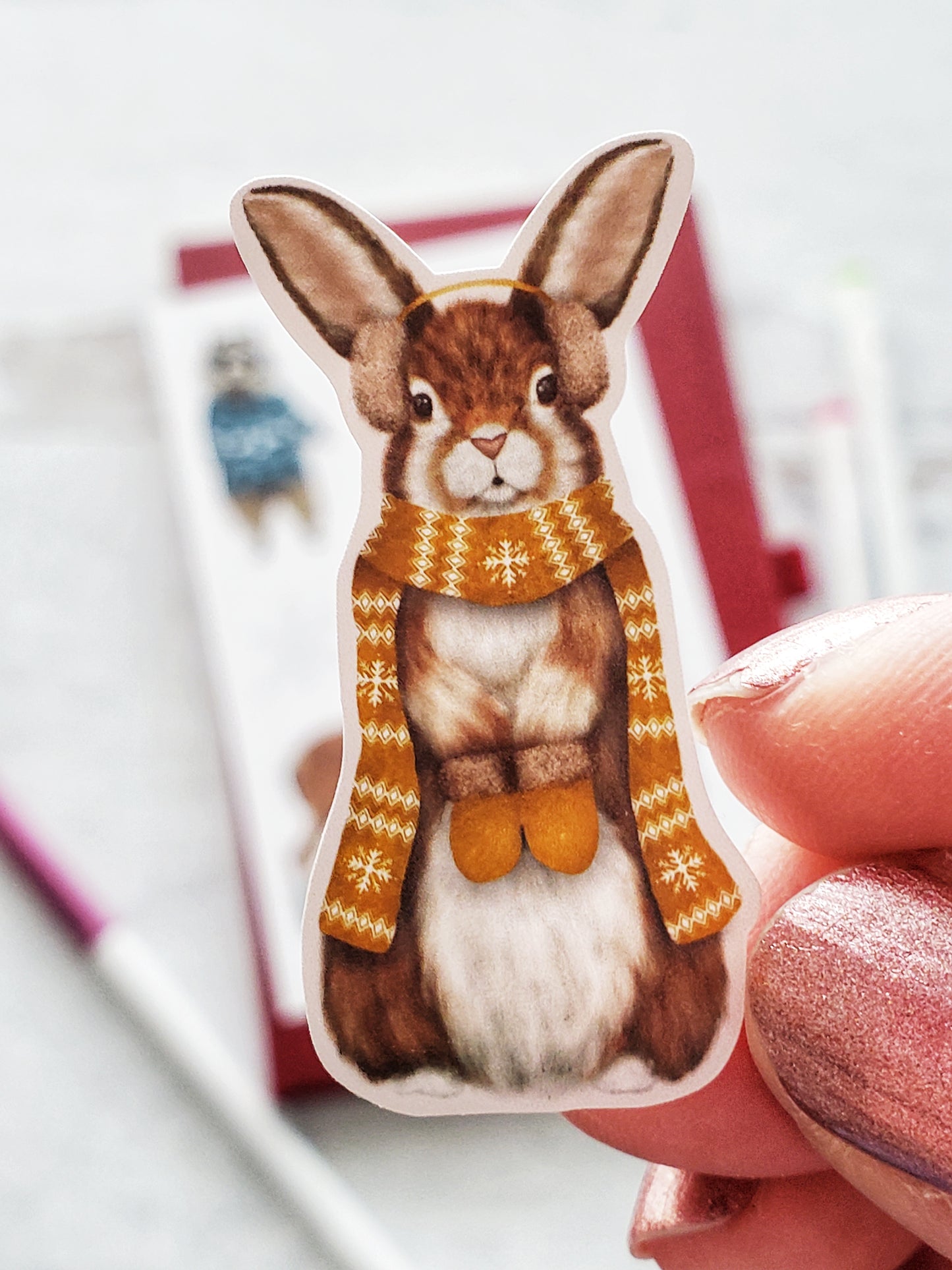 Close up of a sticker of an illustrated rabbit wearing a yellow scarf, mittens and ear muffs.