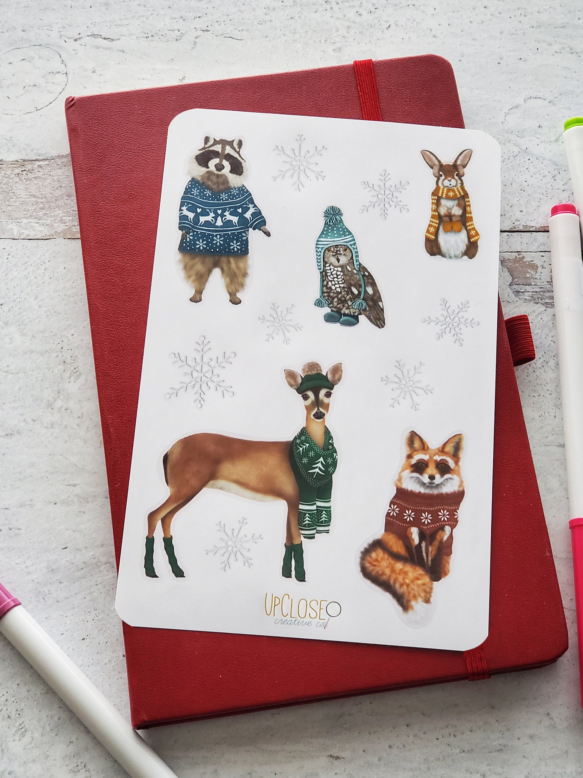 Sticker sheet featuring illustrated forest animals in holiday knits. This sheet includes a racoon, rabbit, owl, deer and fox all wearing colorful sweaters, hats or scarves with festive Christmas prints. 