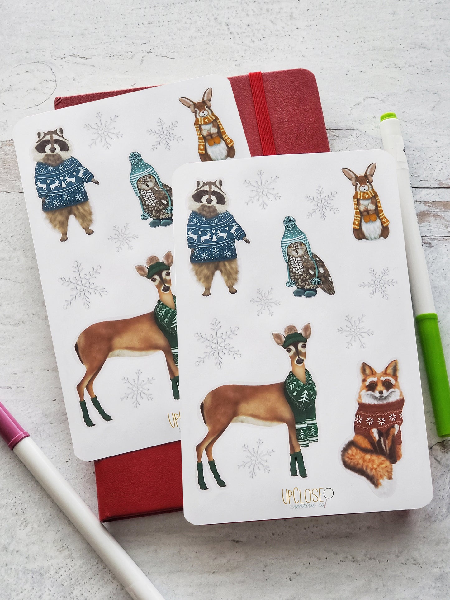 Set of two sticker sheets featuring illustrated forest animals in holiday knits. This sheet includes a racoon, rabbit, owl, deer and fox all wearing colorful sweaters, hats or scarves with festive Christmas prints. 