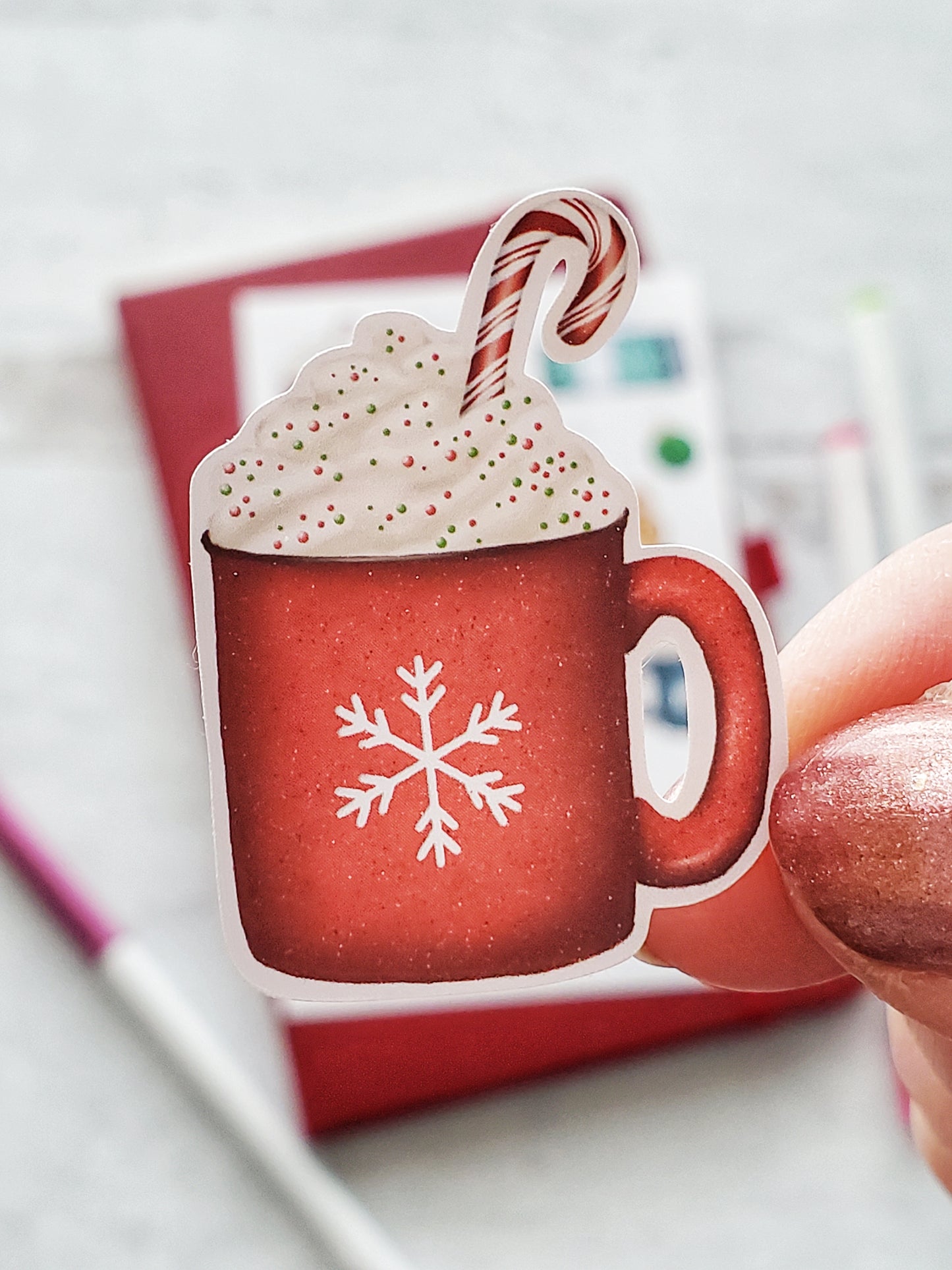 Close up of a sticker of a red hot cocoa mug. The mug has a white snowflake on it and is topped with whipped cream, sprinkles and a red candy cane. 