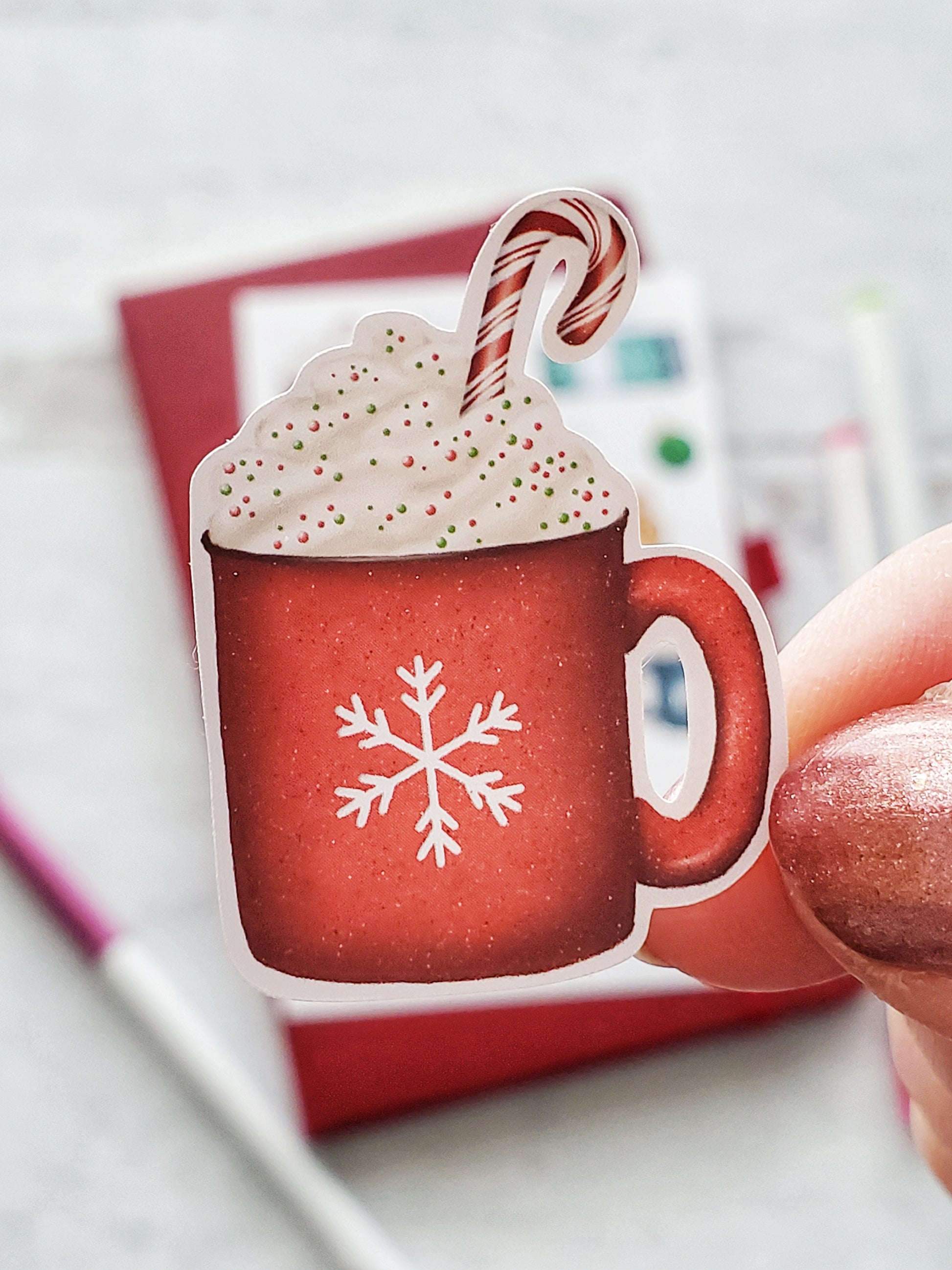 Close up of a sticker of a red hot cocoa mug. The mug has a white snowflake on it and is topped with whipped cream, sprinkles and a red candy cane. 