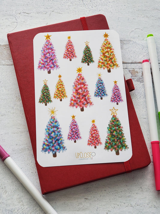 Sticker sheet featuring illustrated Christmas trees of different colors. The trees are purple, pink, green, yellow and blue and each have a different gold topper.