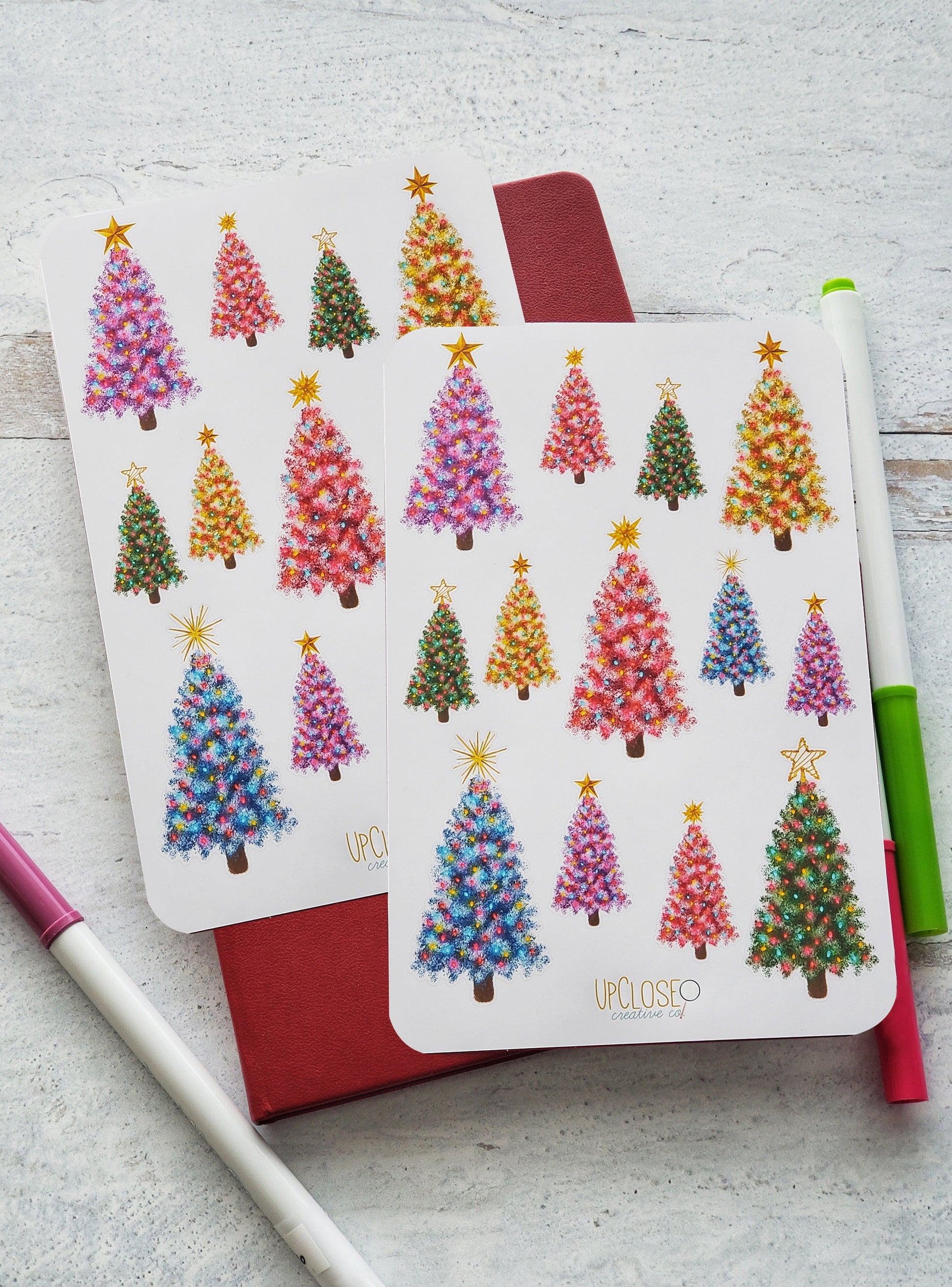 Set of 2 sticker sheets featuring illustrated Christmas trees of different colors. The trees are purple, pink, green, yellow and blue and each have a different gold topper.